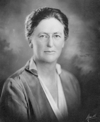 A wearing a simple suit, with wire rim glasses and her hair pulled back.