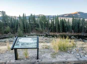 A low exhibit behind a log barrier on a overlook of a river bordered by trees with a mountain in the background