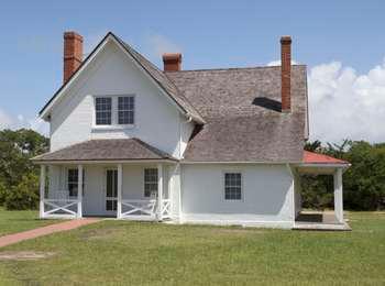 Front view of the white, two-story home with wooden shingles, three red-brick chimneys, and front and side porches.