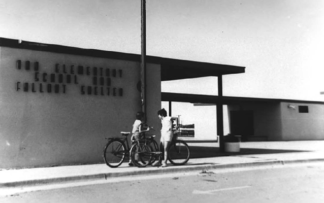 Two children with bicycles stand in front of square structure with flagpole.