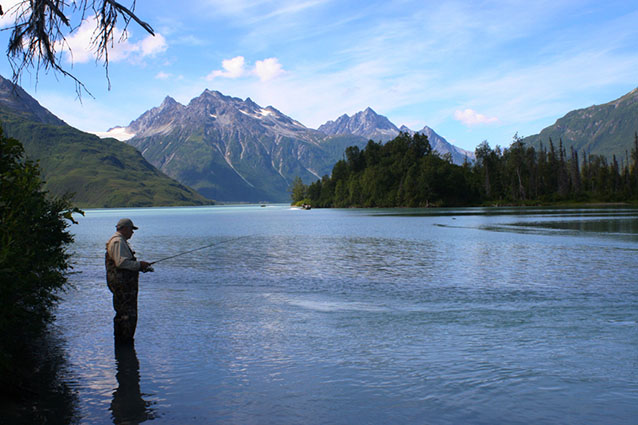 Photo of a man fishing in a lake surrounded by forests and tall, rocky mountains.