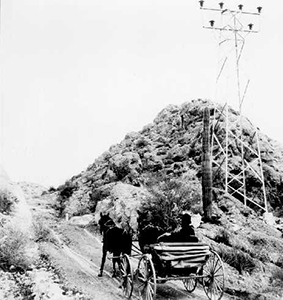 Black and white photo of driver and a horse drawn carriage next to tall metal towers 