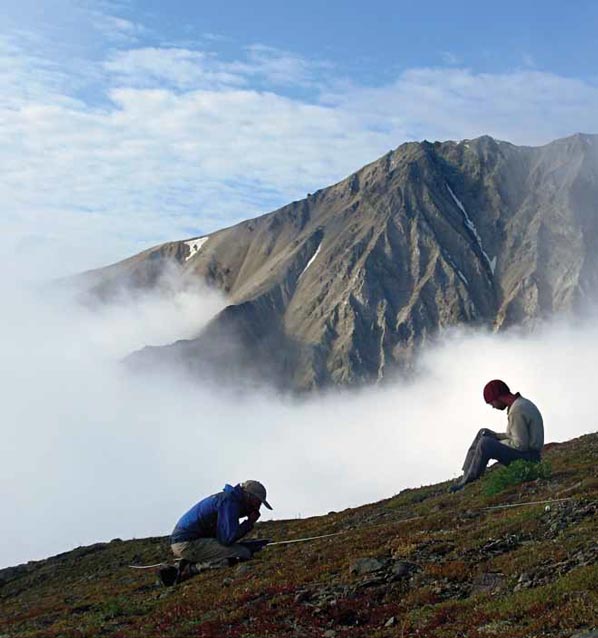 two people sitting on a hillside overlooking a distant mountain shrouded in clouds