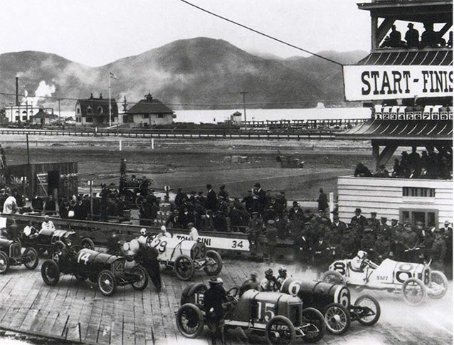 Onlookers gather near Grand Prix cars on a wooden track beside the start/finish line, 1915.