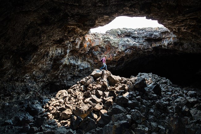 Caver walks over a pile of rocks beneath a sky light in a cave.