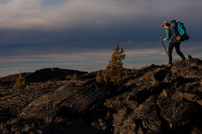 Woman with backpack and hiking poles traverses over rocky lava.