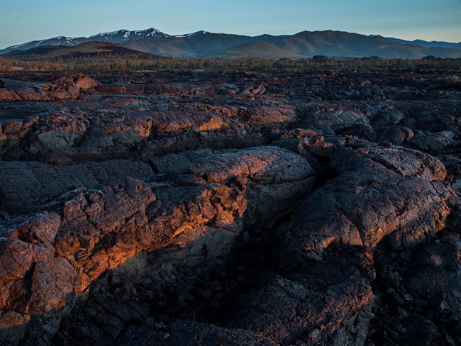 Ropey, pahoehoe lave stretches along the ground in front of snow-capped mountains.