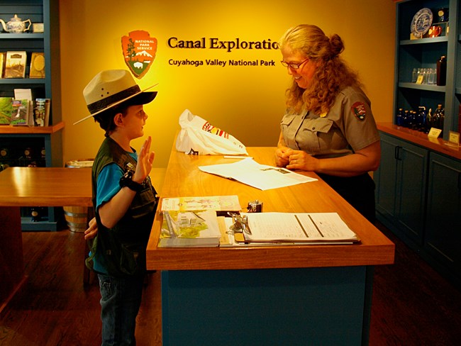 A junior ranger stands at the desk holding their right hand up and wearing a ranger hat; behind the desk, a ranger checks their work