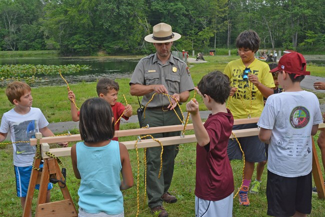Uniformed ranger and several children stand outside along two boards with yellow ropes in their hands.