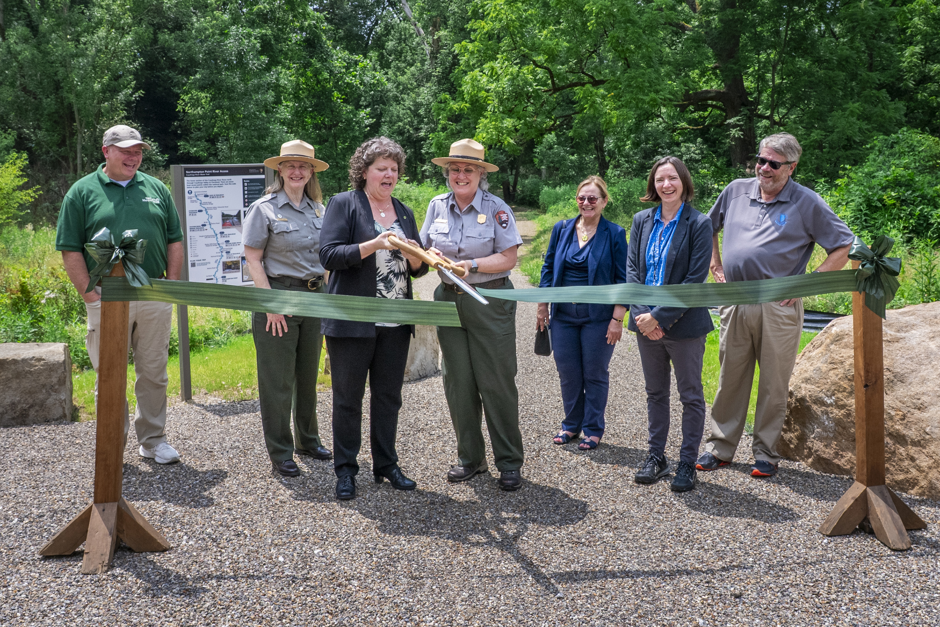 A group of seven people, some uniformed rangers, stand smiling behind a green ribbon across a trail, while two of them cut that ribbon with oversized scissors.