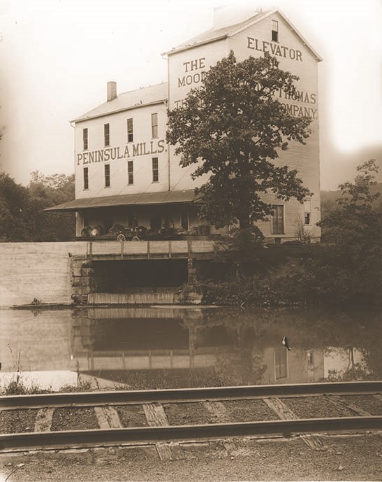 Black-and-white photo of a three-story white building and its reflection in a body of water; railroad tracks in the foreground; words on the building partially obscured by a tree include the words “Peninsula Mills”.