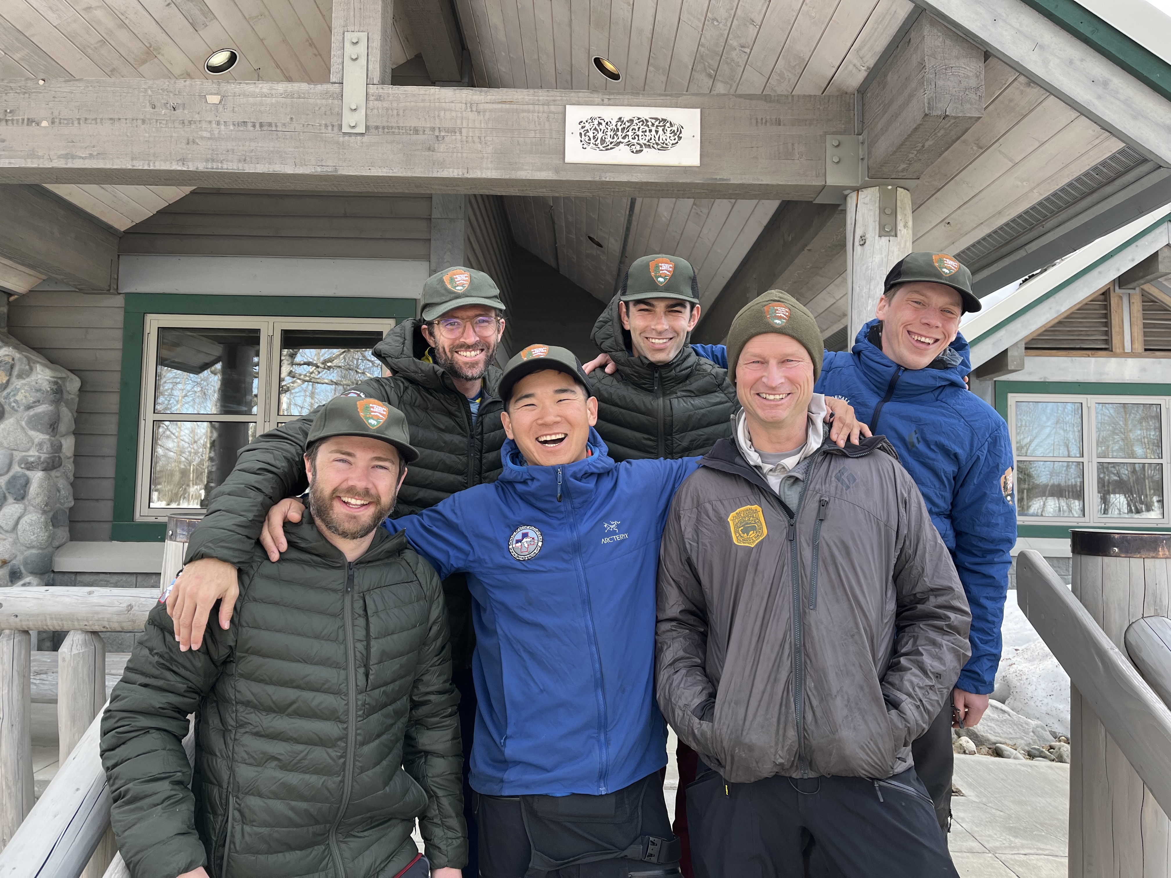 Six smiling men in winter clothing stand on steps outside a ranger station