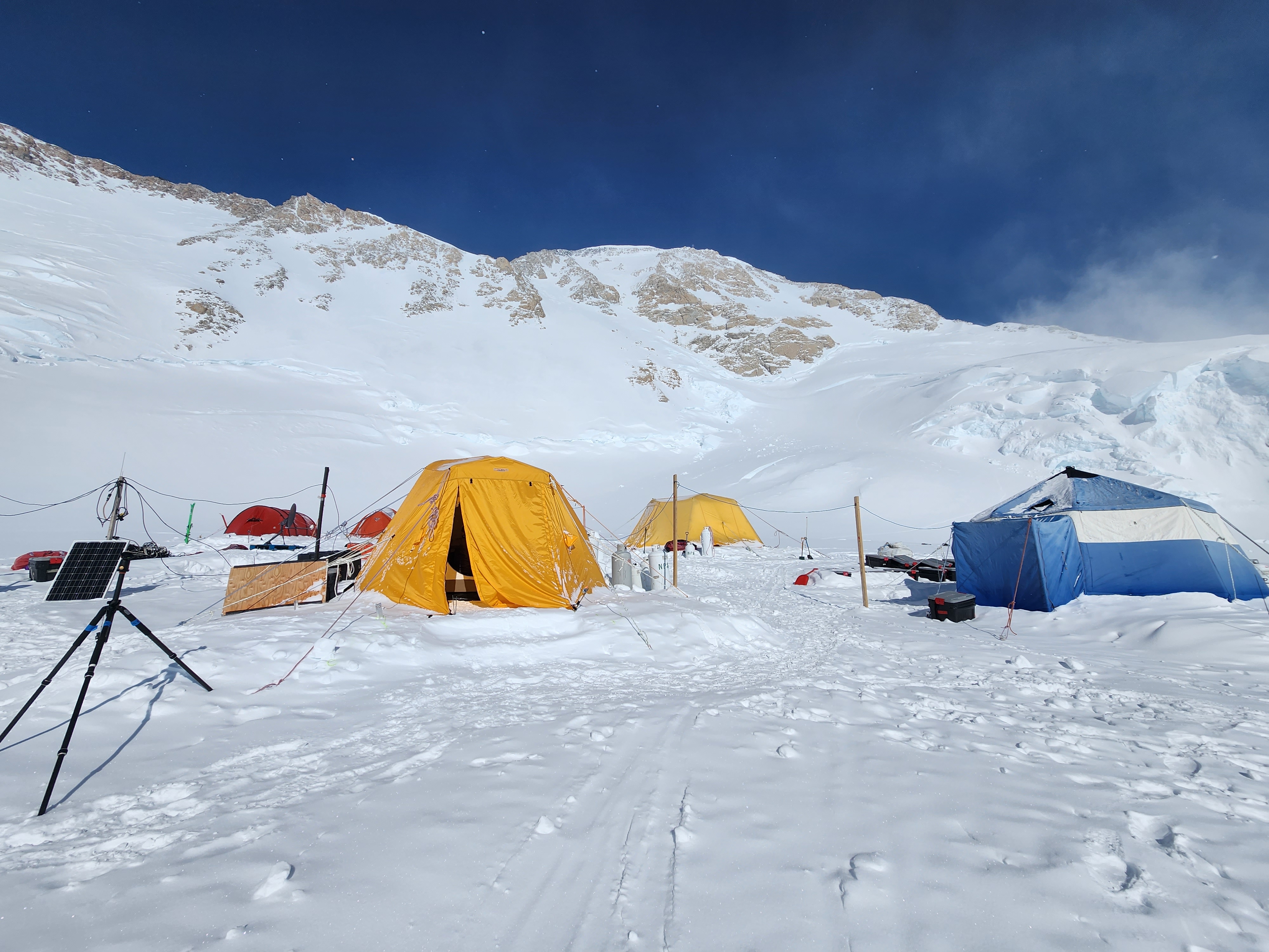 Glacier occupied by five large camp tents, several of which are connected to a solar panel with electrical wires.