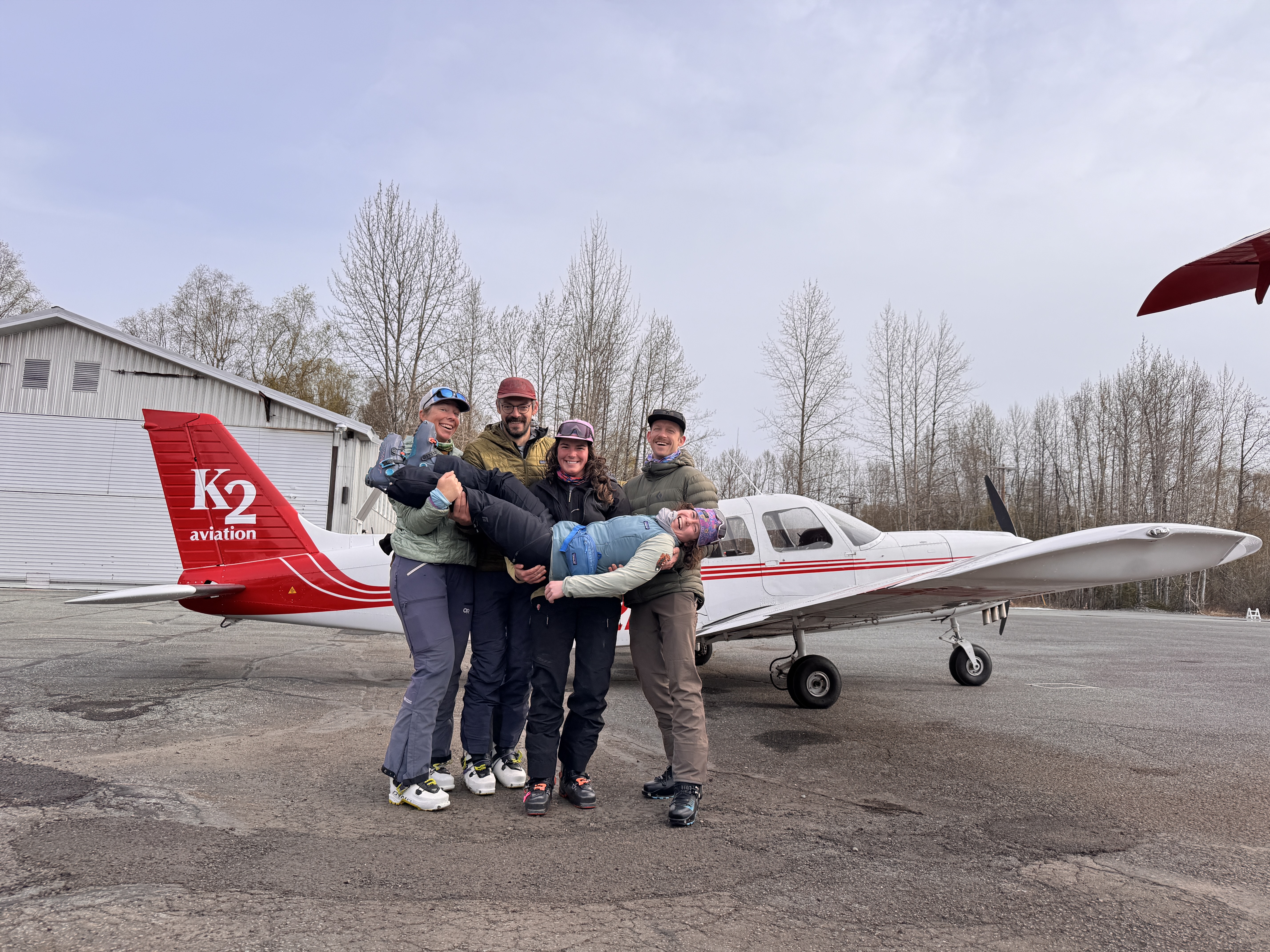 4 people stand in a line holding up a fifth person in front of a red and white plane