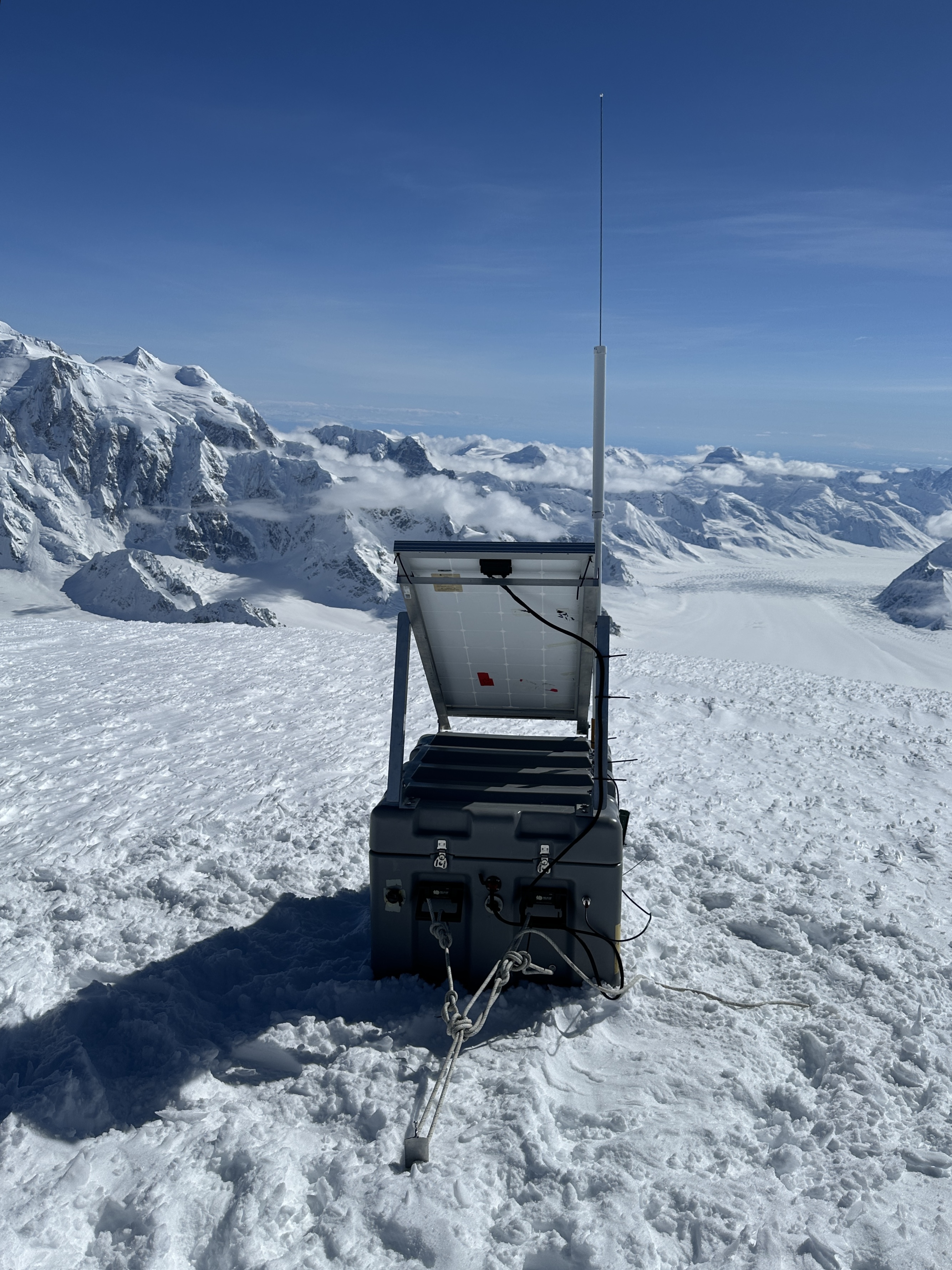 A big black box with a solar panel and antenna is sectured to a snow field with ropes