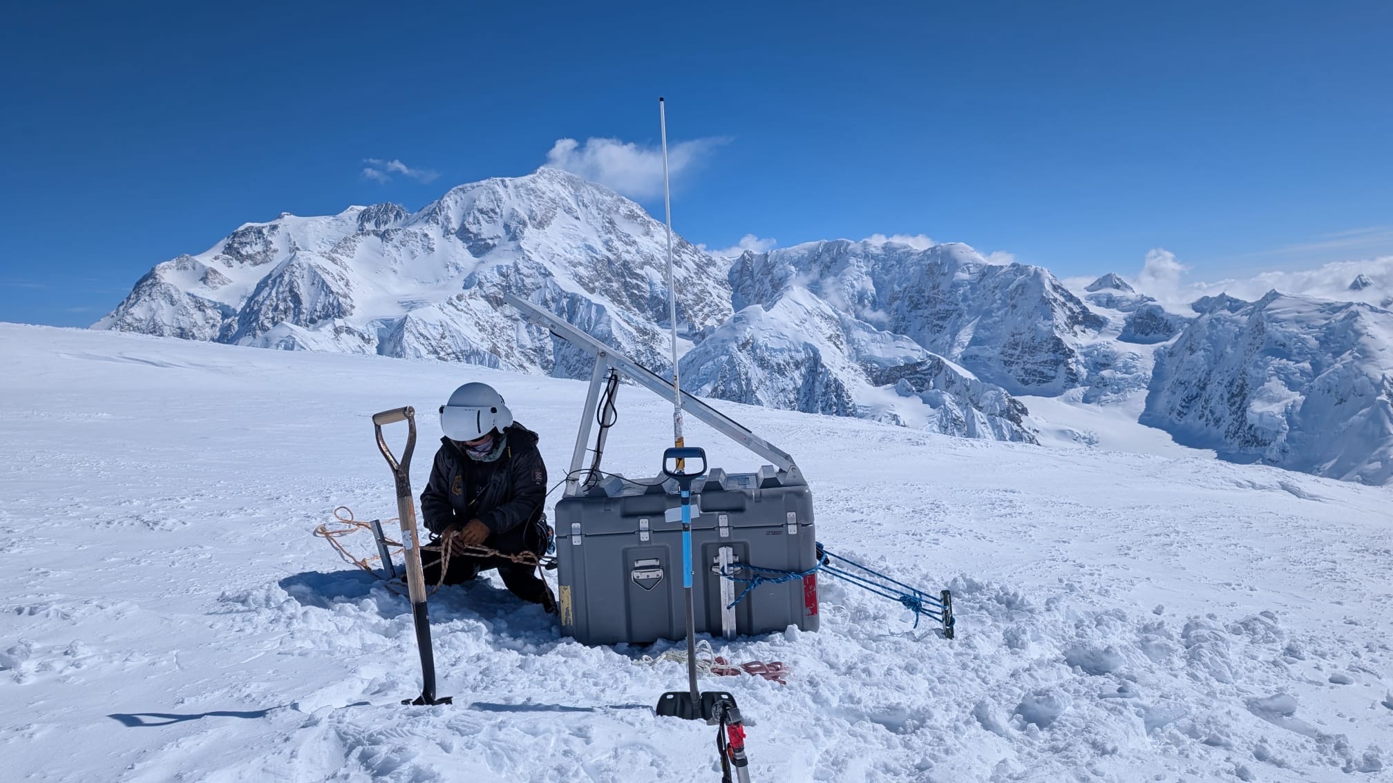 A ranger wearing a helmet works on a large grey device surrounded by snowy peaks