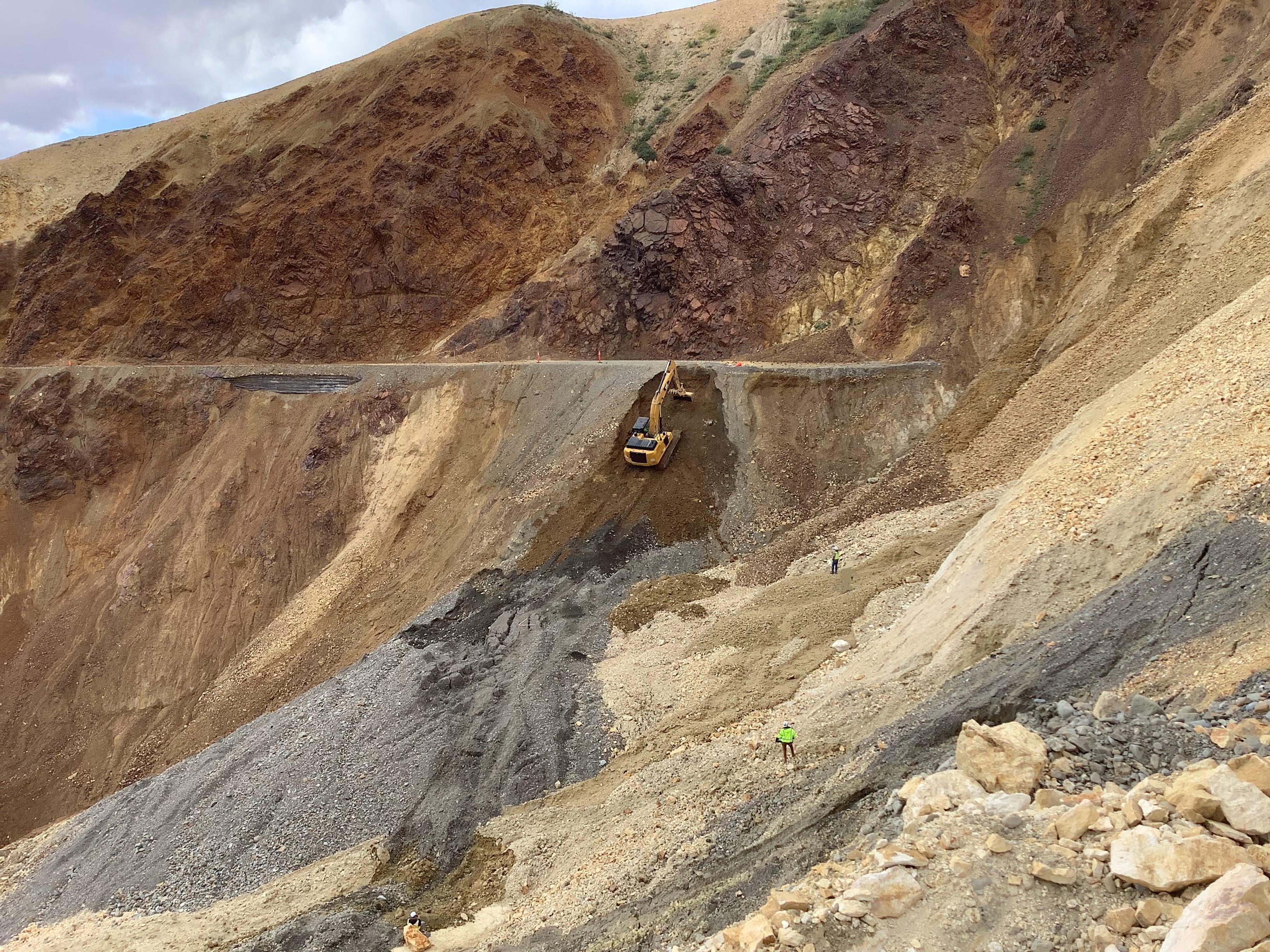 An excavator climbs up a steep slope to the flat road on the far side of the Pretty Rocks landslide. Two people in safety vests observe.
