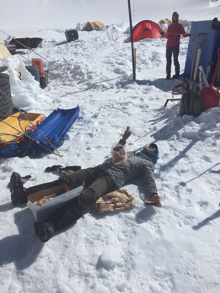 Mountaineer lies flat in the snow with a box of donuts and tub of ice cream on her chest