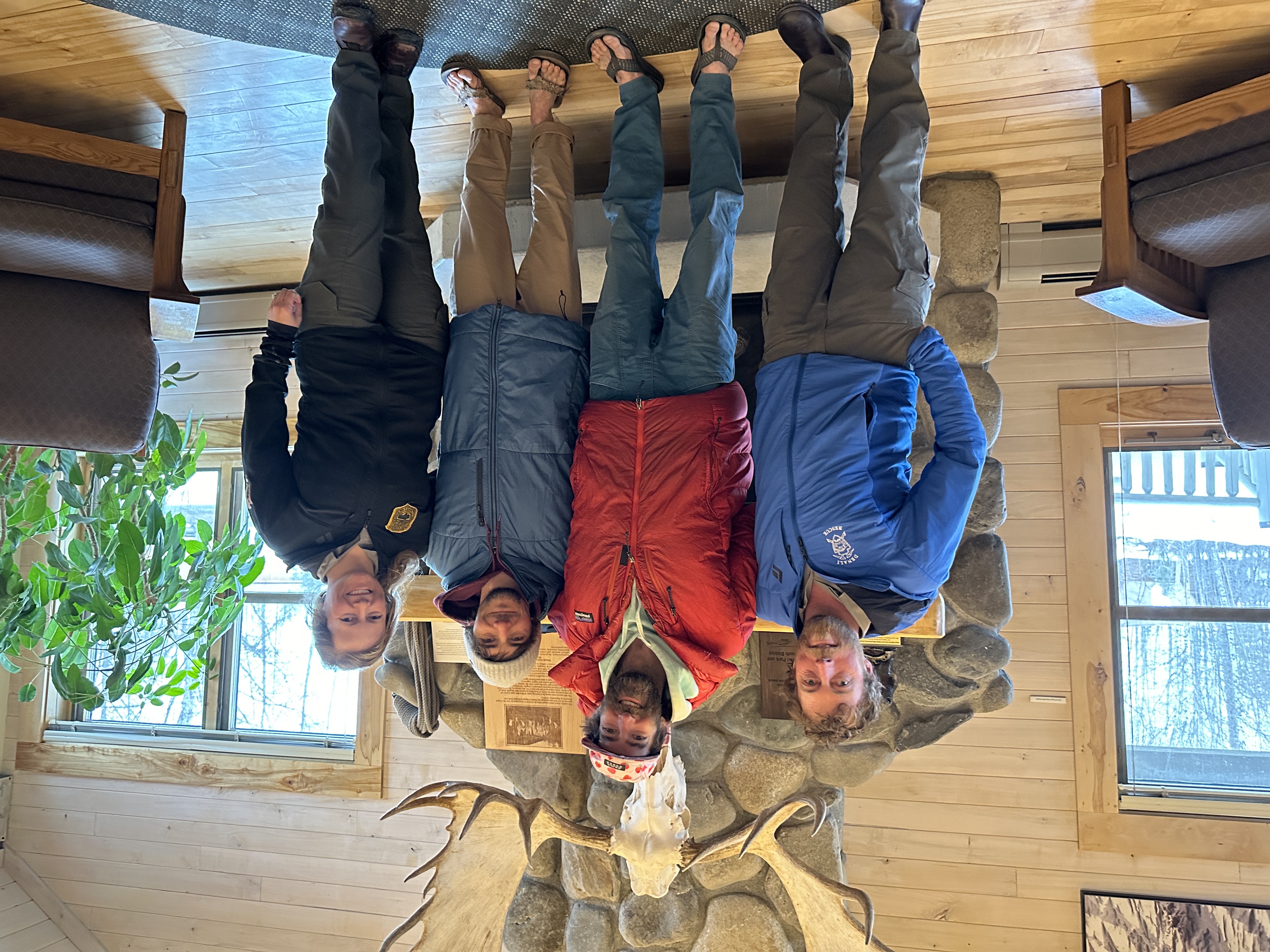 Two rangers and two climbers in puffy jackets and flip-flops pose in front of the ranger station fireplace