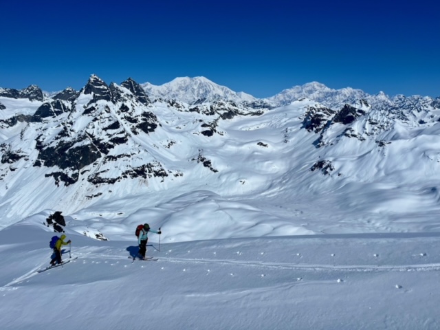 Two skiers with backpacks follow ski tracks along a snowfield
