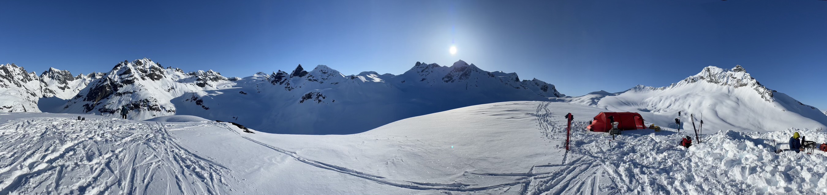 Panoramic image of a mountain range with a tent camp and many ski tracks in the snow