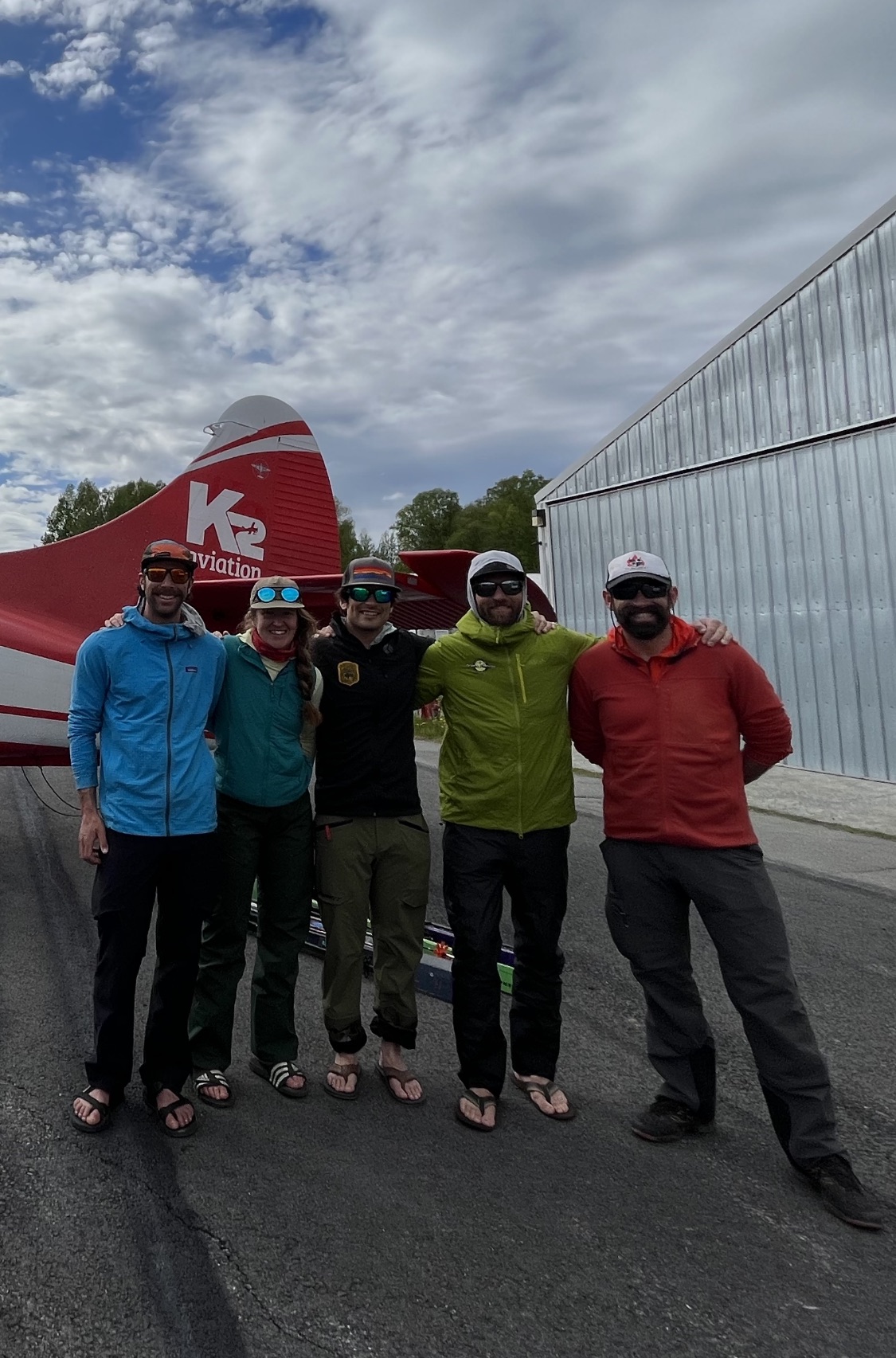 Five climbers in flip flops stand on an airport runway in front of a small aircraft