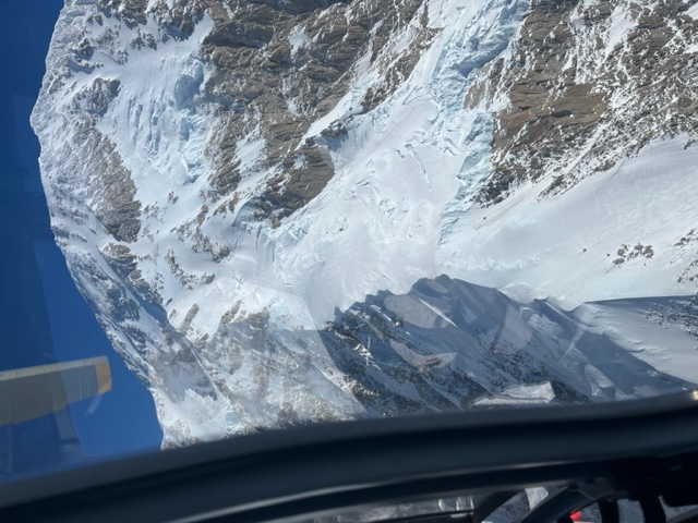 Image looking out the window of a helicopter down at a snow and ice covered mountain ridge