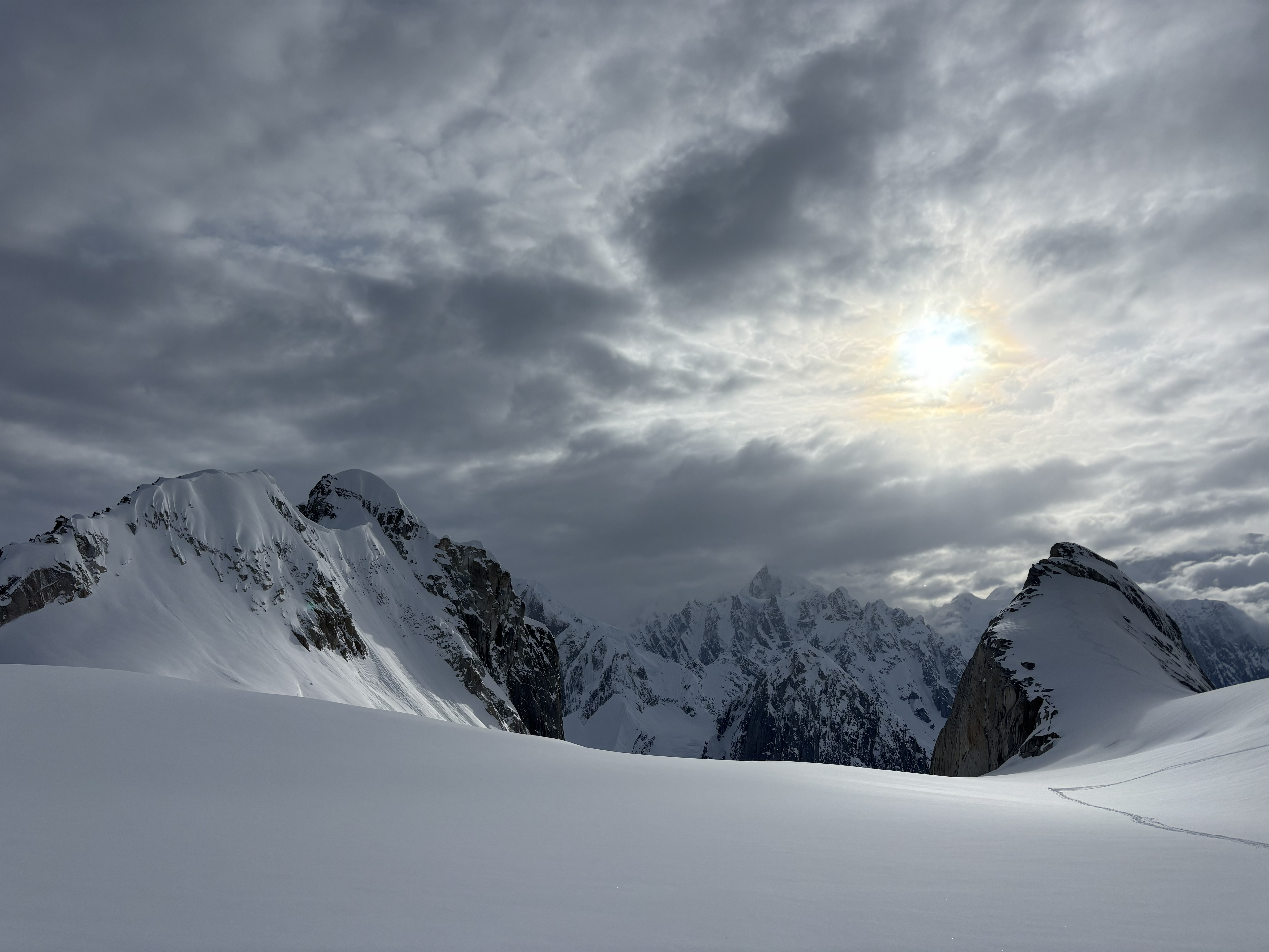 Snow covered mountain peaks in front of a grey cloudy sky with the sun shining