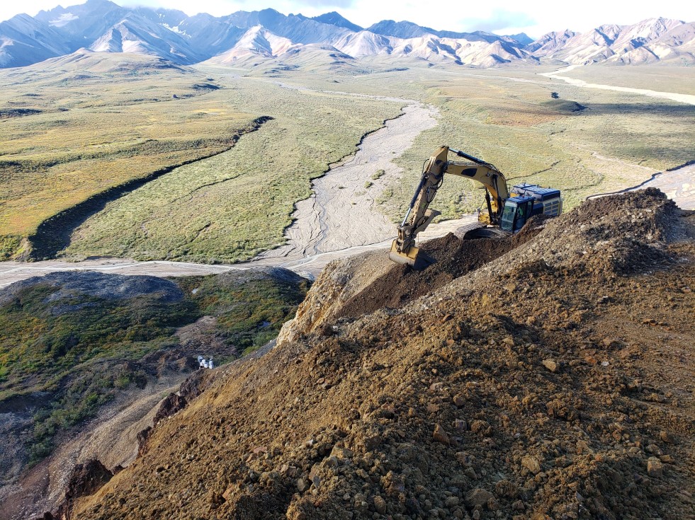 Looking down a steep slope to an excavator moving rocks. Below, the flat Plains of Murie at the base of the slope stretch out toward the mountains of the Alaska Range.