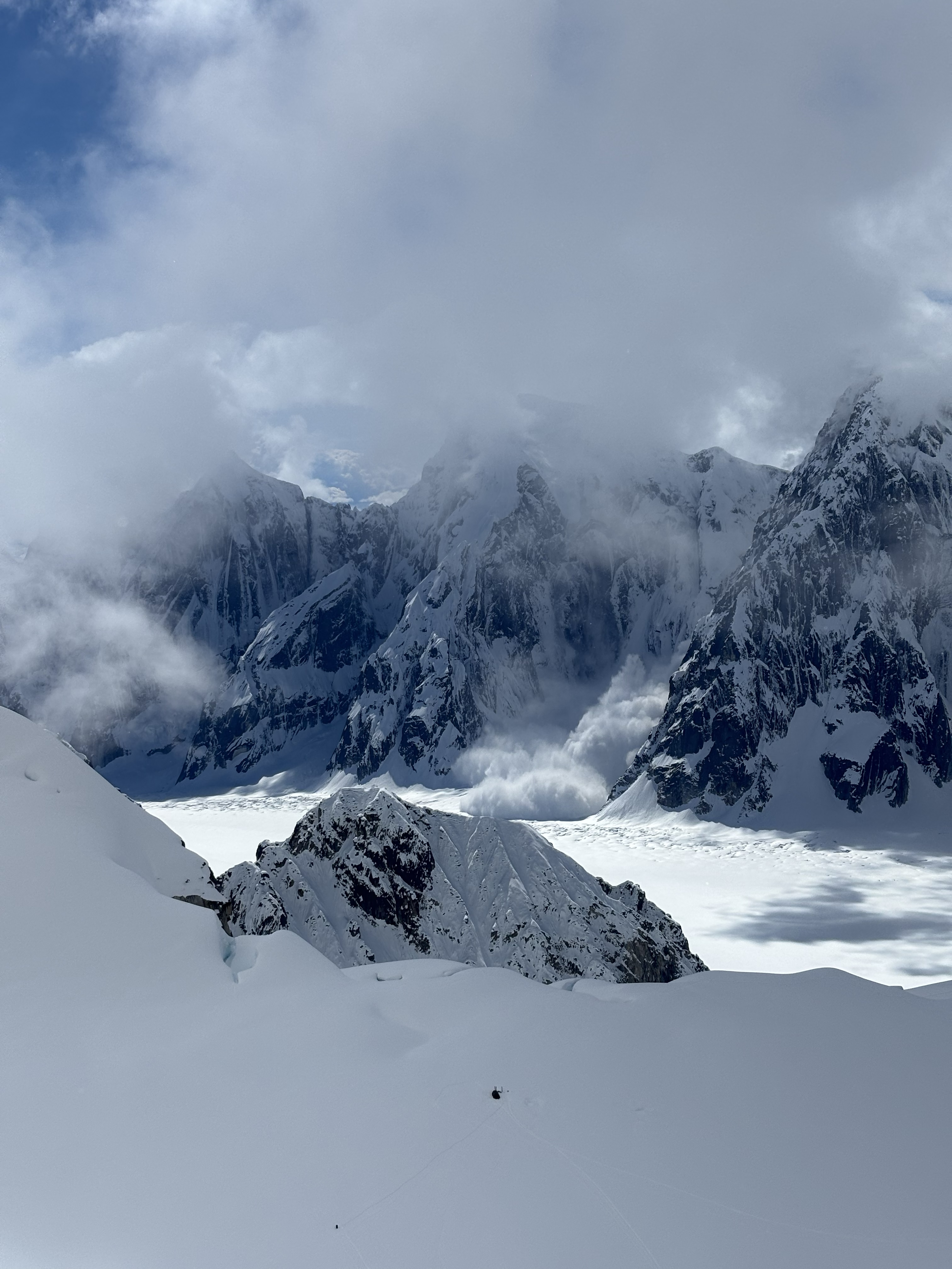 Grey snowy peaks with a wall of snow sliding down to a snow-covered field with a large group of clouds above.