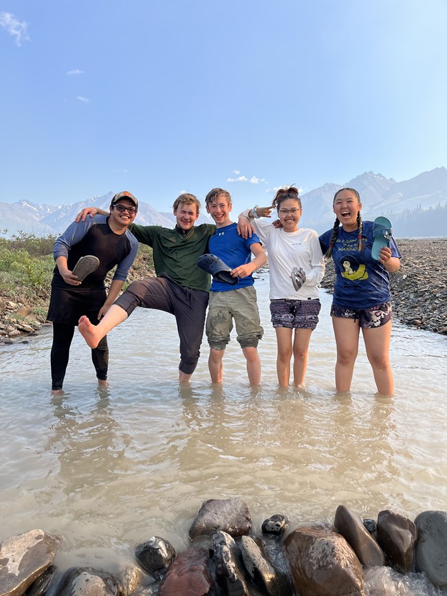 5 people wade in a river holding their shoes and posing.