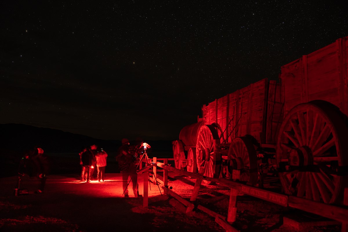 Night image. Red lights illuminate people a camera on a tripod a wooden fence and historic twenty mule team wagons.