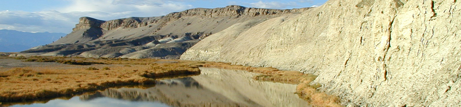 The image captures a vast, serene landscape featuring a calm river that reflects the surrounding rugged terrain. Massive cliffs dominate the right side while rolling mountains stretch across the horizon under a partially cloudy sky.