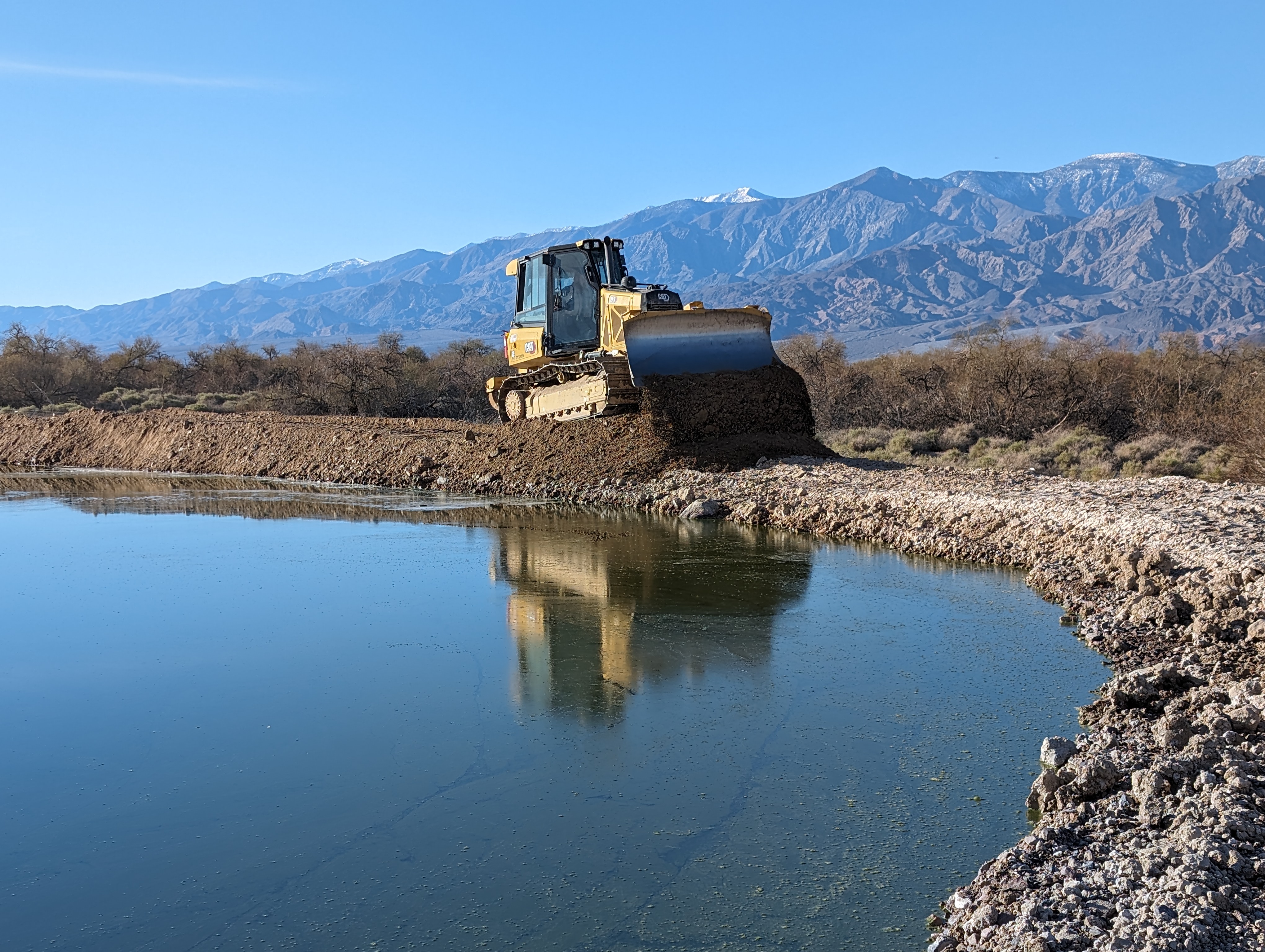 A yellow bulldozer pushes dirt on the berm of a pond of blue water.