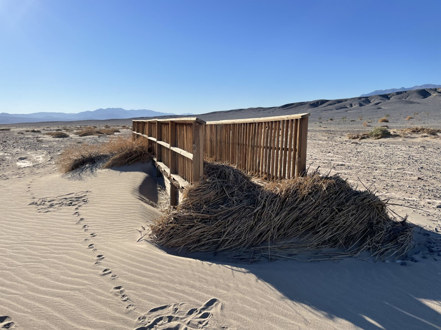 Boardwalk bridge overtaken with sand and brush with mountains in the background.