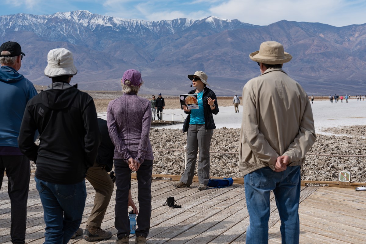 A woman wearing a sun hat, holding a photo talks to a group of people while standing on a wooden boardwalk with salt flats and snow-capped mountains in the background.