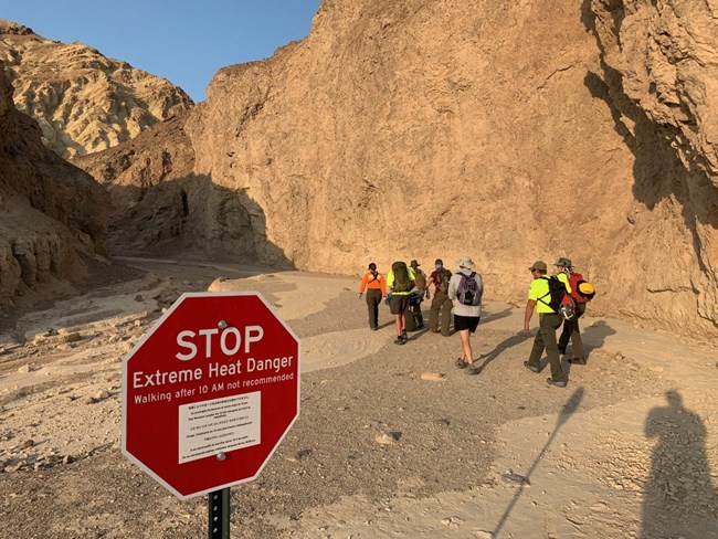 Red, octogen shaped sign warning visitors of heat danger. Behind the sign, a group of park rangers hike into a canyon with backpacks and other rescue gear.