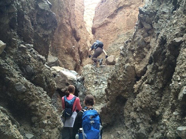 A hiker climbs a dryfall inside a narrow canyon while two other hikers prepare for their turns.