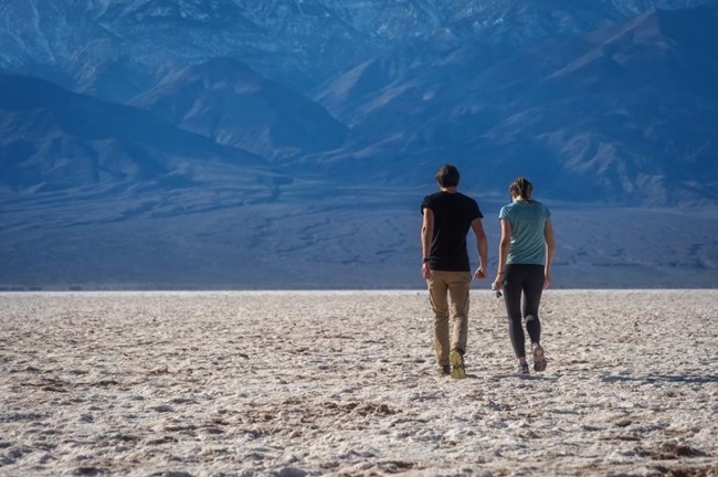 A man and woman walk across salt flats toward large mountains.