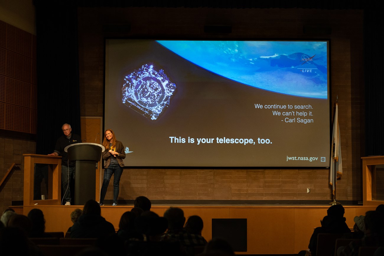 A man and woman stand near a podium on a stage with a large screen in the background which reads: This is your telescope, too.