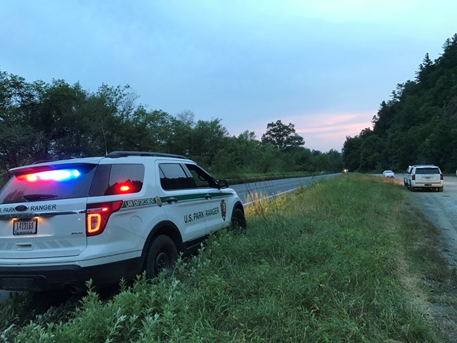 A park ranger patrol vehicle with its lights on pulled on the grass next to a road.