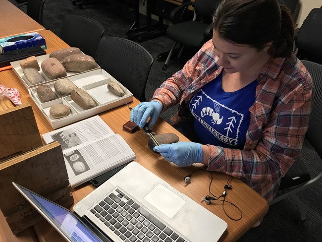 A person with blue gloves on sits at a table surrounded by rocks, books, and a laptop. They are using a tool to measure the width of the rock.