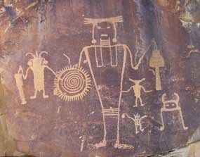 Large petroglyph of human-like figure with trapezoidal body and with several other human-like figures around it.