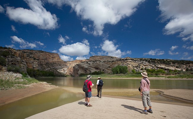 Three hikers stand beside the Yampa River looking at a rocky ridge.