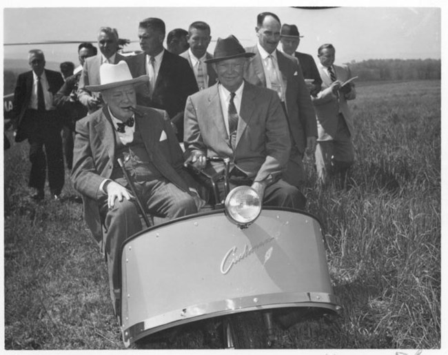 Winston Churchill and Dwight Eisenhower tour the Eisenhower in a tiny car. A group of men stand in the background.