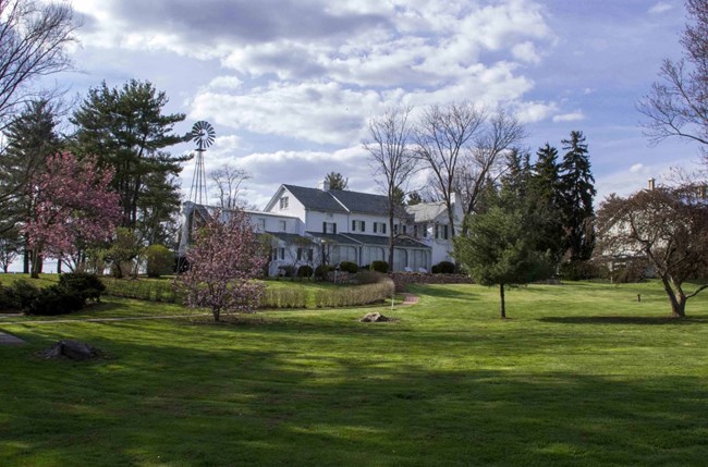 The large white brick home belonging to Dwight and Mamie Eisenhower stands in the background. In front is green grass and red blooming trees in springtime.