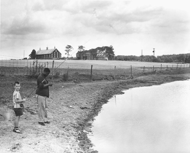 A young David Eisenhower stands with John Moaney on the banks of a pond on the Eisenhower property; both are holding fishing rods. A home stands in the background.