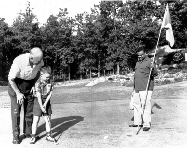President Eisenhower helps Grandson David put a golf ball; John Moaney is standing at the hole, holding a flag