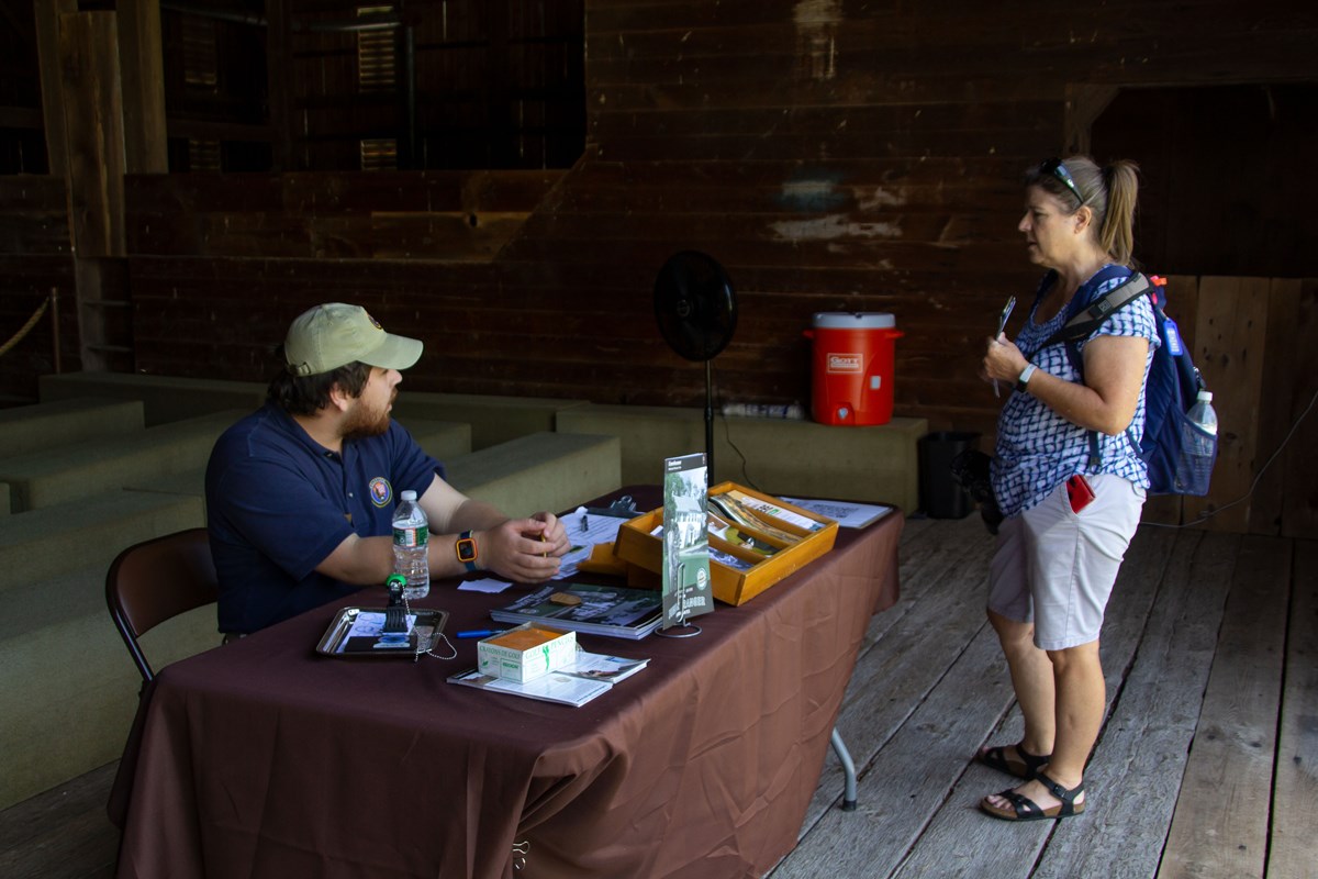 A park visitor speaks with an intern at an information table in a barn.