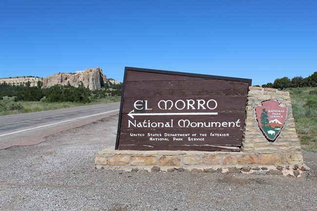 A sign reading "El Morro National Monument" with a large rock cliff in the background.